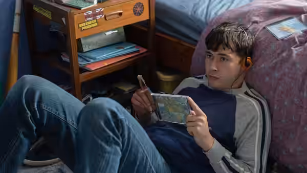 A young man reclines beside a bed, holding a map and listening through earbuds