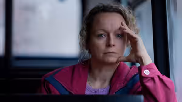 A woman in a pink jacket rests her hand against her forehead indoors