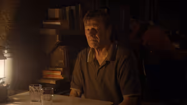 Jem Stoker (Sean Bean) sits at a table beside a lamp and drinking glasses