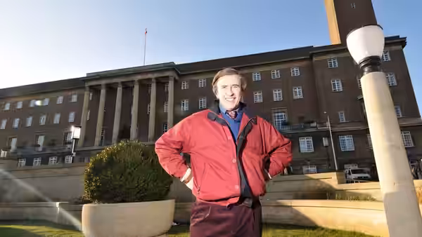 Alan Partridge (Steve Coogan) stands with his hands on his hips outside a large institutional building