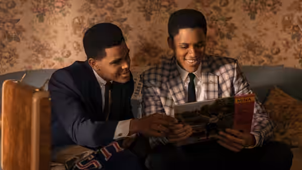 Two young men smile as they read a magazine together on a sofa