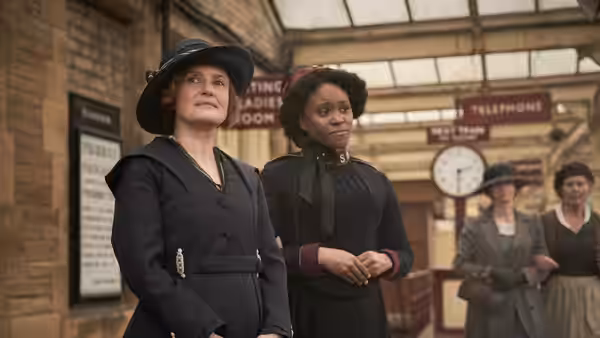 Mary Lockwood (Amara Okereke) stands beside a woman in a hat inside a railway station