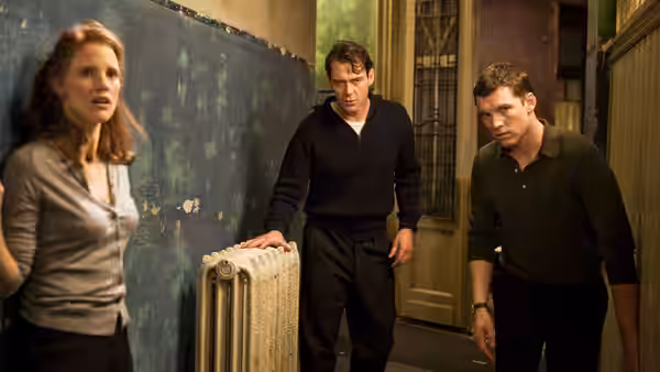 A woman stands beside a radiator as two men enter a worn, narrow room