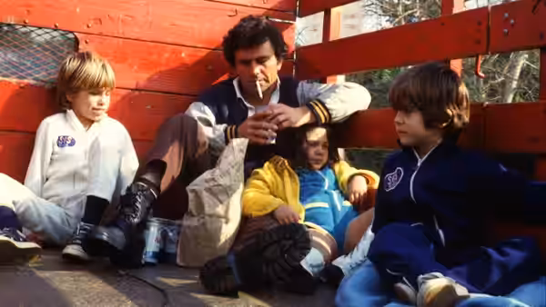 Nick Longhetti (Peter Falk) sits with children in matching uniforms against a red wall