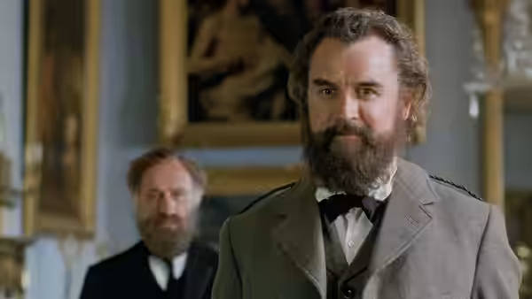 A bearded man in a grey suit stands in a palace room, with another man behind him