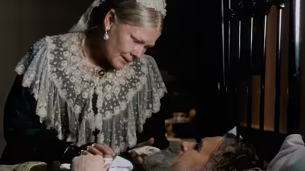 Queen Victoria (Judi Dench) leans over a bed and holds the hand of a reclining figure