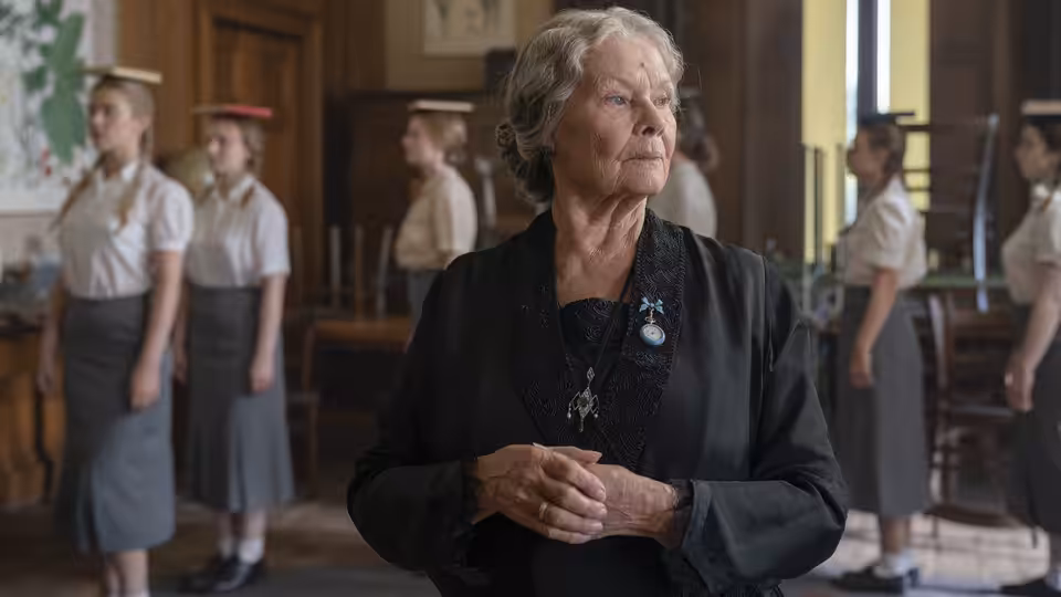 Miss Rocholl (Judi Dench) stands among uniformed students inside a school hall