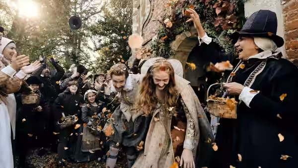 A bride and groom bow as guests shower them with petals outside a house