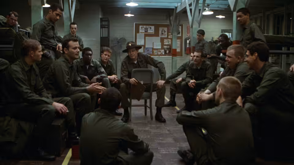 Sgt. Hulka (Warren Oates) sits among recruits gathered in a barracks room