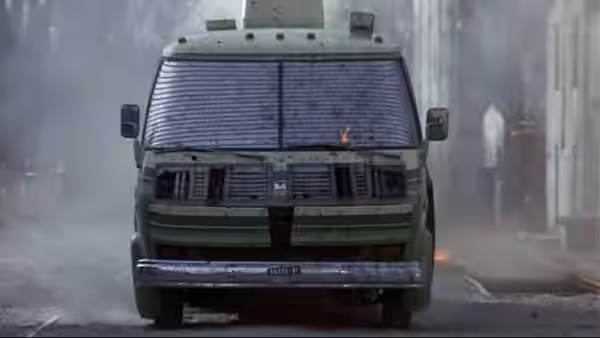 A military van drives through smoke with flames flickering on its windshield