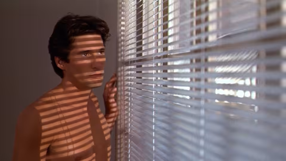 Julian Kaye (Richard Gere) peers through venetian blinds, striped sunlight falling across his bare chest