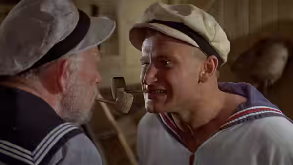 Popeye (Robin Williams) leans toward an elderly sailor in a wooden room