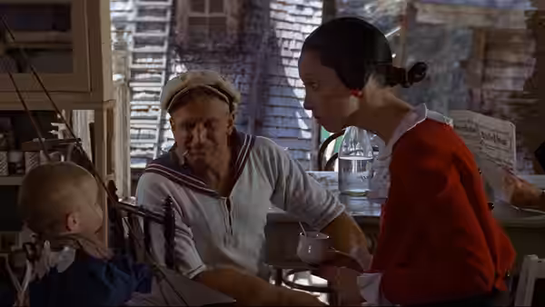 Popeye (Robin Williams) and Olive Oyl (Shelley Duvall) sit with their baby at a table