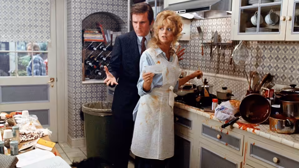 Glenda Parks (Goldie Hawn) cooks in a cluttered kitchen as Nick Gardenia (Chevy Chase) stands behind her