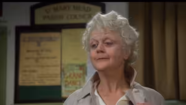 A white-haired woman stands before a parish council noticeboard