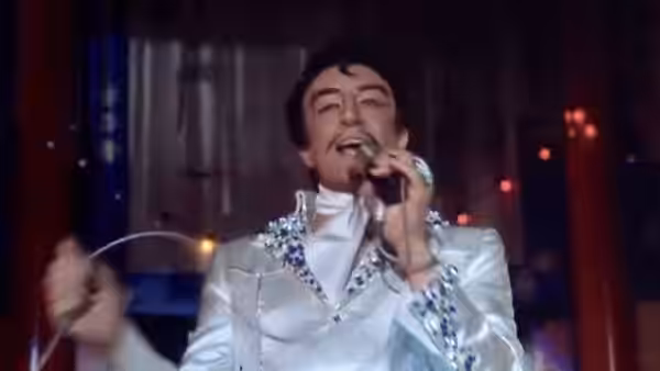 A dark-haired performer sings into a microphone, wearing a jeweled white suit