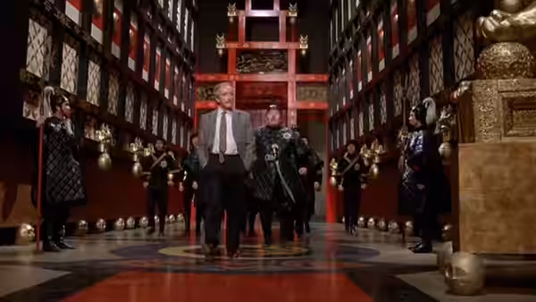 Fu Manchu (Peter Sellers) leads a gray-haired man through a ceremonial hall lined with guards