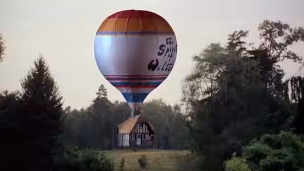 A striped hot-air balloon hovers above a small house in a wooded clearing