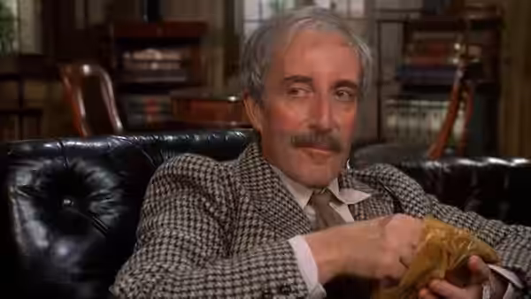 Sir Roger Avery (David Tomlinson) sits on a leather sofa holding a paper bag