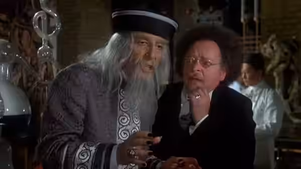 Fu Manchu (Peter Sellers) leans close to a bespectacled man in a laboratory