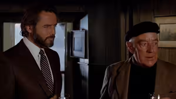 A bearded man stands beside an elderly man wearing a black beret indoors