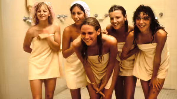 Five young women in towels gather beneath showerheads in a locker room