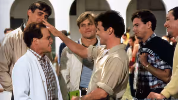 A young man touches his friend's forehead while several teammates watch in a school courtyard