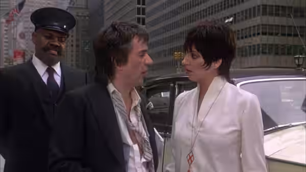Arthur Bach (Dudley Moore) talks with Linda Marolla (Liza Minnelli) beside a car as Bitterman watches