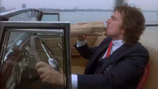 Arthur Bach (Dudley Moore) drives a convertible and drinks from a paper bag beside the water