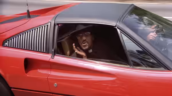 A moustached man waves from the open window of a red sports car