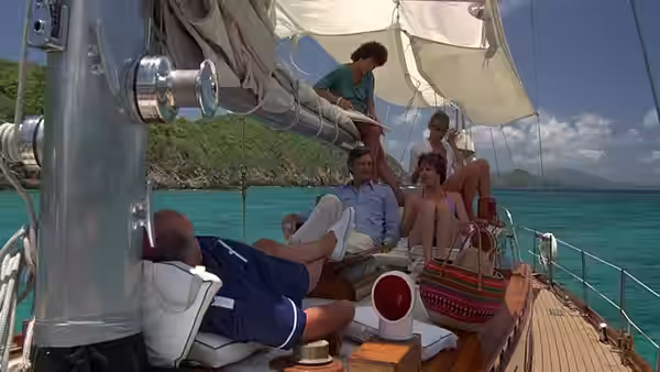 Four friends relax aboard a sailboat near a green, rocky shoreline