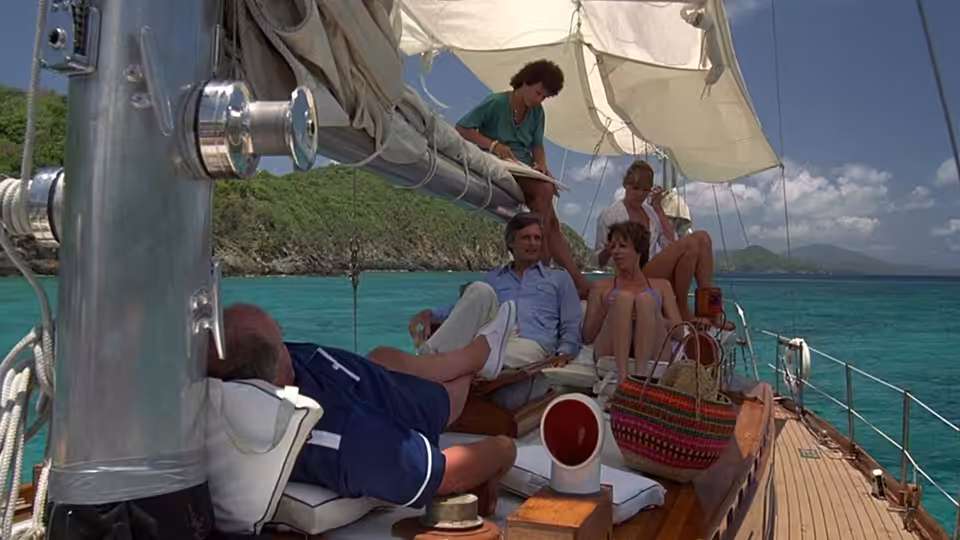 Four friends relax aboard a sailboat near a green, rocky shoreline