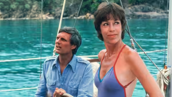 A man and woman stand aboard a sailboat on turquoise water