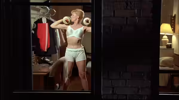 A blonde woman lifts dumbbells beside an open wardrobe in a bedroom