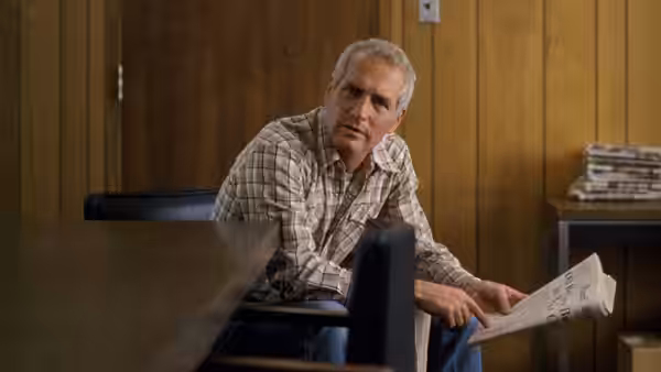 Michael Colin Gallagher (Paul Newman) sits holding a newspaper in a wood-paneled room