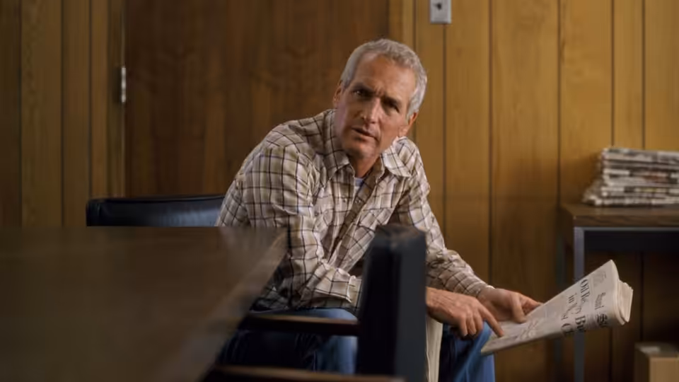 Michael Colin Gallagher (Paul Newman) sits holding a newspaper in a wood-paneled room
