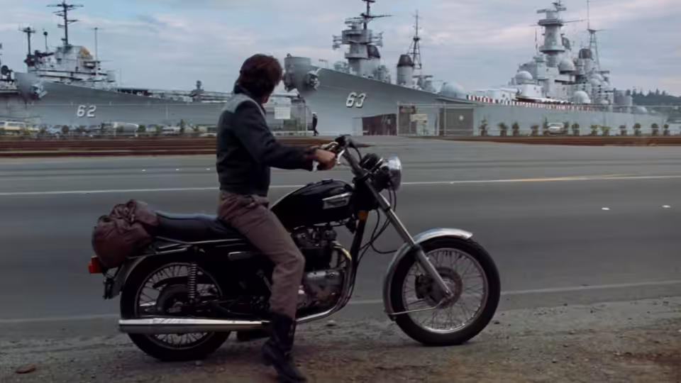 Zack Mayo (Richard Gere) sits astride his motorcycle beside docked Navy ships