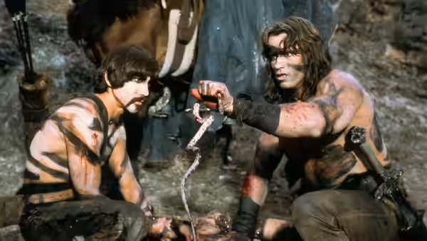 Conan (Arnold Schwarzenegger) and Subotai (Gerry Lopez) crouch together, examining a snake