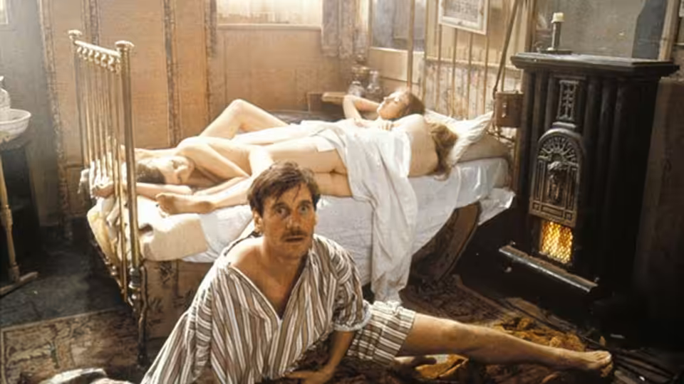 The Reverend Charles Fortescue (Michael Palin) sits on the floor beside women sprawled across a bed