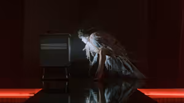 A dancer crouches beside a television on a dark stage lit by red floor lights