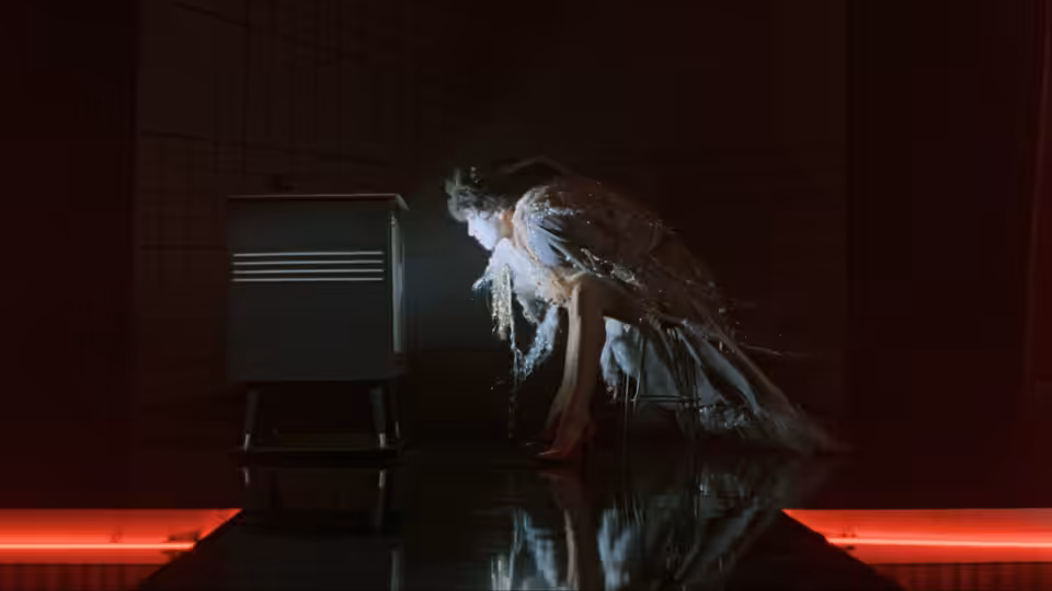 A dancer crouches beside a television on a dark stage lit by red floor lights