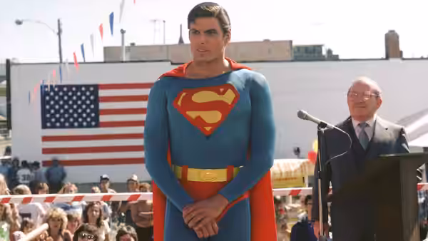 Superman (Christopher Reeve) stands before an American flag as a man speaks at a microphone