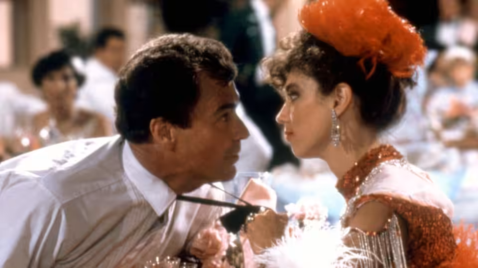 A man leans close to a woman in a feathered red hat at a crowded dance