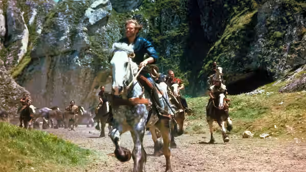 Prince Colwyn (Kenneth Marshall) rides a white horse through a rocky forest with mounted companions