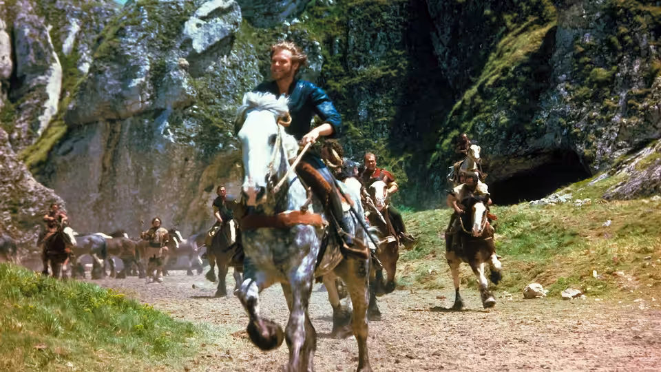 Prince Colwyn (Kenneth Marshall) rides a white horse through a rocky forest with mounted companions