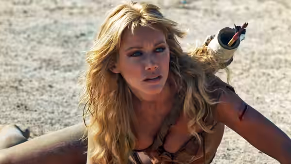 Sheena crouches in the sand with an arrow lodged near her shoulder