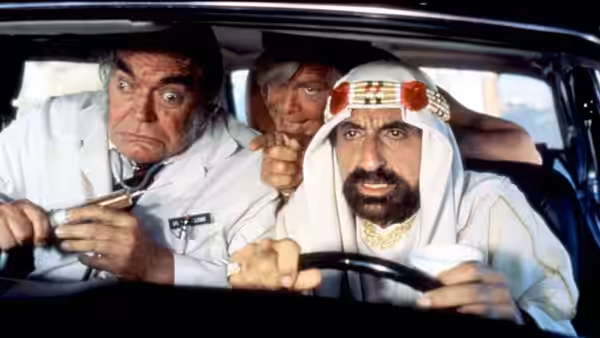 A bearded man in a white robe grips the steering wheel as two men crowd into the car
