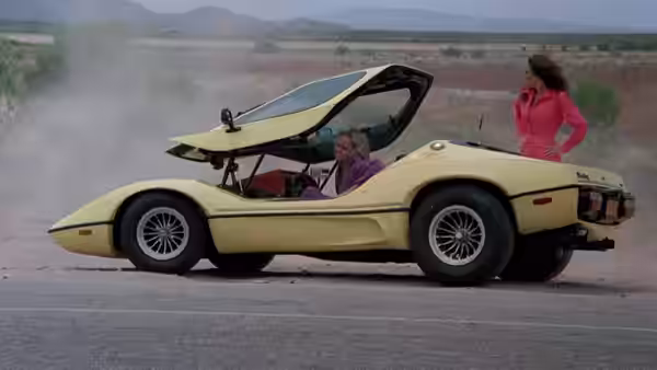 A woman sits in a yellow sports car while another stands beside it on the roadside