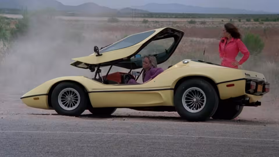 A woman sits in a yellow sports car while another stands beside it on the roadside