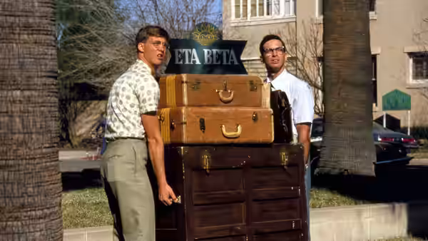 Gilbert Lowell (Anthony Edwards) and Lewis Skolnick (Robert Carradine) carry luggage outside the Eta Beta house